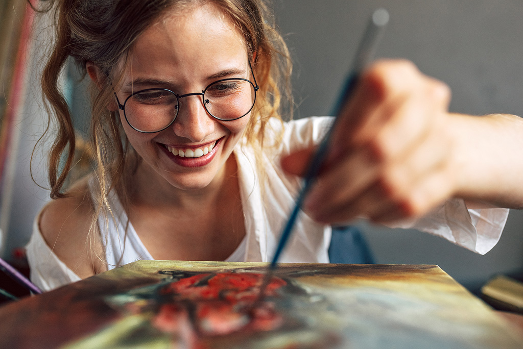 een lachende jonge vrouw met een grote bril schildert.
